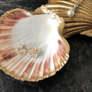 Puede incluir: Un par de pendientes de oro con detalles de cristal transparente se muestran en una concha blanca con borde dorado. La concha est&aacute; sobre una superficie gris oscura.