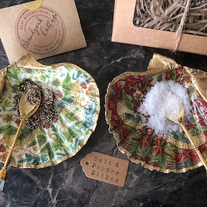 Könnte beinhalten: Zwei goldgeränderte Keramik-Salz- und Pfefferstreuer in Muschelform mit Blumenmustern. Jedes Schälchen hat einen goldenen Löffel mit einem klaren Kristallgriff und ist mit Salz und Pfeffer gefüllt.