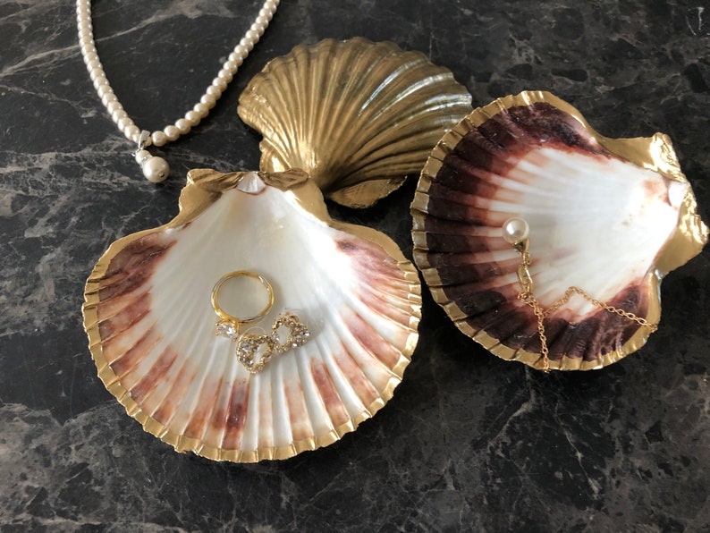 Puede incluir: Dos conchas decorativas con detalles dorados, una con una perla y una cadena, la otra con un anillo y dos peque&ntilde;os amuletos de piedras preciosas.