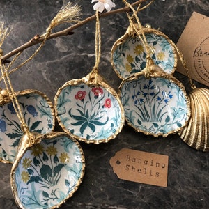 May include: Six gold-painted seashells with floral patterns, hanging from a branch. The shells are decorated with blue, red, and yellow flowers. The text "Hanging Shells" is on a brown tag.