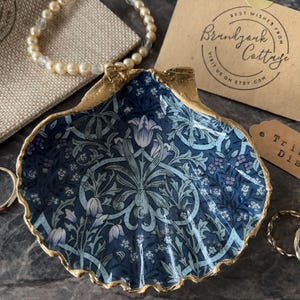May include: A decorative seashell trinket dish featuring a dark blue and light blue floral pattern with gold-painted edges. The dish is accompanied by a beige burlap pouch, a pearl bracelet, two gold rings, a branded card, and a tag labeled Trinket Dish.
