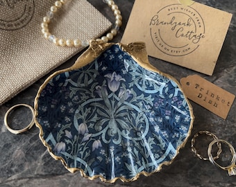 Puede incluir: Un plato decorativo de concha marina con un patrón floral en azul oscuro y azul claro con bordes pintados en oro. El plato se acompaña de una bolsa de arpillera beige, una pulsera de perlas, dos anillos dorados, una tarjeta y una etiqueta.