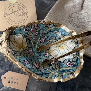 May include: A decorative sugar bowl with a floral pattern in teal, cream, and pink, edged with gold. Includes gold-toned tongs, a burlap bag, and a tag that reads "Sugar Bowl". A card with the text "Best Wishes From Brandjank Cottage" is also present.