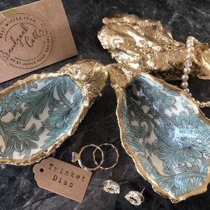 May include: Two gold oyster shell trinket dishes with a blue and white floral pattern inside. The dishes are open and a pearl necklace, two rings, and a pair of earrings are inside. A brown tag with the text "Trinket Dish" is in the lower left corner.