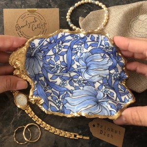 May include: A gold trinket dish with a blue and white floral design. The dish is shaped like a shell and has a gold rim. The text "Trinket Dish" is written on a brown tag.