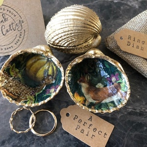 May include: Two gold seashell ring dishes with a woodland scene inside. The shells are open and the dishes are sitting on a grey surface. There is a small brown tag that says "A Perfect Pair" and a larger brown tag that says "Ring Dish".
