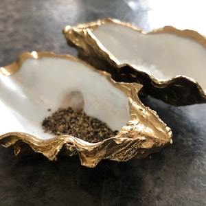 Op de afbeelding: Twee met goud geverfde oesterschelpen, een gevuld met zwarte peperkorrels. De schelpen zijn een decoratief keukenaccessoire.
