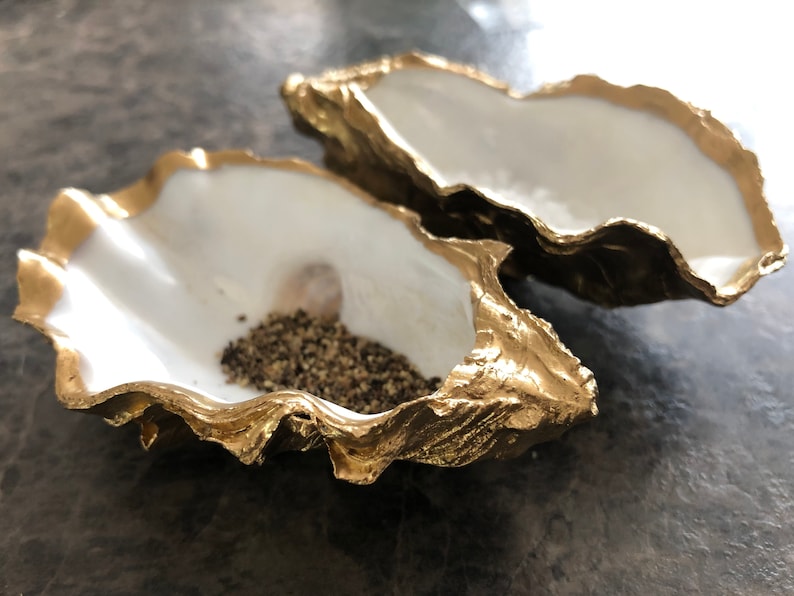 Op de afbeelding: Twee met goud geverfde oesterschelpen, een gevuld met zwarte peperkorrels. De schelpen zijn een decoratief keukenaccessoire.