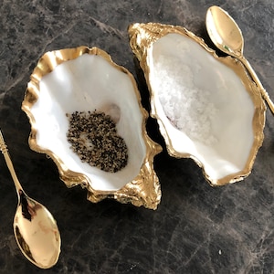 Op de afbeelding: Twee gouden gerande oesterschelpen, een gevuld met zwarte peperkorrels en de andere met wit zout, met twee gouden lepels met heldere kristallen handvatten.