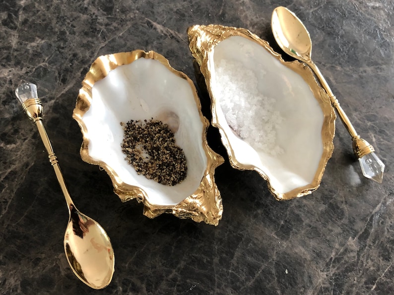 Op de afbeelding: Twee gouden gerande oesterschelpen, een gevuld met zwarte peperkorrels en de andere met wit zout, met twee gouden lepels met heldere kristallen handvatten.