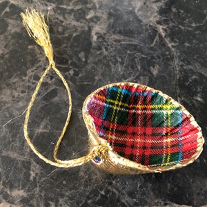 May include: A gold-colored seashell ornament with a red, green, blue, and yellow plaid fabric interior. The ornament has a gold string for hanging. The seashell is a decorative item.