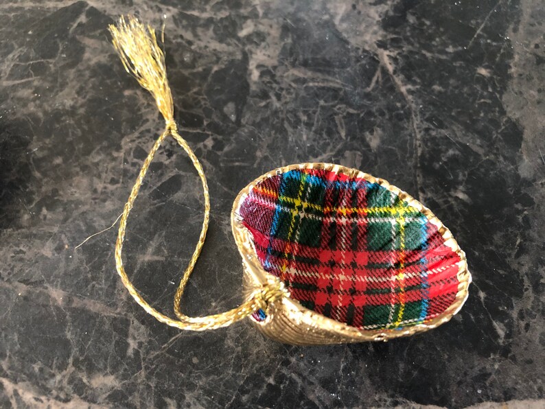May include: A gold-colored seashell ornament with a red, green, blue, and yellow plaid fabric interior. The ornament has a gold string for hanging. The seashell is a decorative item.