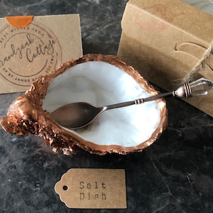 May include: A decorative salt dish made from a natural oyster shell, with a copper-colored rim and a white interior. A small spoon with a crystal handle rests inside. A tag reads "Salt Dish".