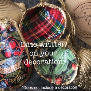 May include: Three gold-painted seashells with different patterned paper inserts. The shells are decorated with the year 2023 and are strung with gold cord. The text "Date written on your decoration" is visible in the image.  The text "Best Wishes from Brandyark Cottage" is visible on a brown paper tag.