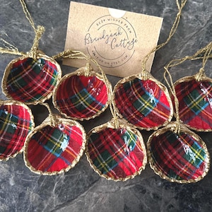 May include: Eight seashell ornaments with a red, green, and blue plaid pattern. Each shell has a gold-colored trim and a gold string for hanging. A small card with text is visible in the background.