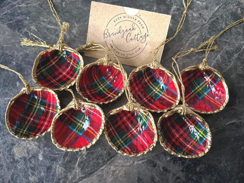 May include: Eight seashell ornaments with a red, green, and blue plaid pattern. Each shell has a gold-colored trim and a gold string for hanging. A small card with text is visible in the background.
