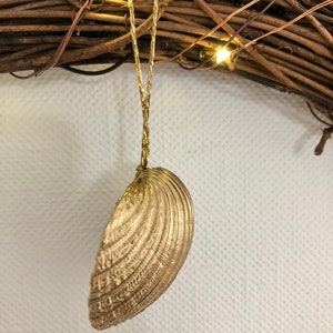 May include: A gold-colored seashell ornament hanging from a gold string. The seashell has detailed ridges and is suspended in front of a brown twig wreath with small, warm white lights.