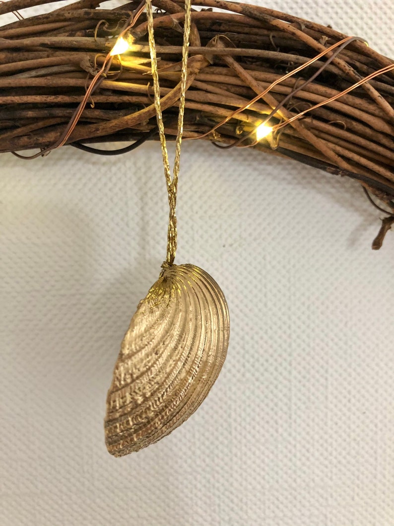 May include: A gold-colored seashell ornament hanging from a gold string. The seashell has detailed ridges and is suspended in front of a brown twig wreath with small, warm white lights.