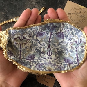 May include: A decorative trinket dish with a gold-painted edge and a floral pattern in shades of purple, blue, and green. The dish is held in open hands. A small tag reads "Brindjack Cotty".