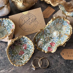 May include: Three decorative oyster shells with gold-painted edges and interior floral decoupage patterns featuring tulips and lilies. One shell contains a single white pearl. Two metal rings are placed nearby, alongside a card labeled Brandyjunk Cottage and a tag marked Ring Box.