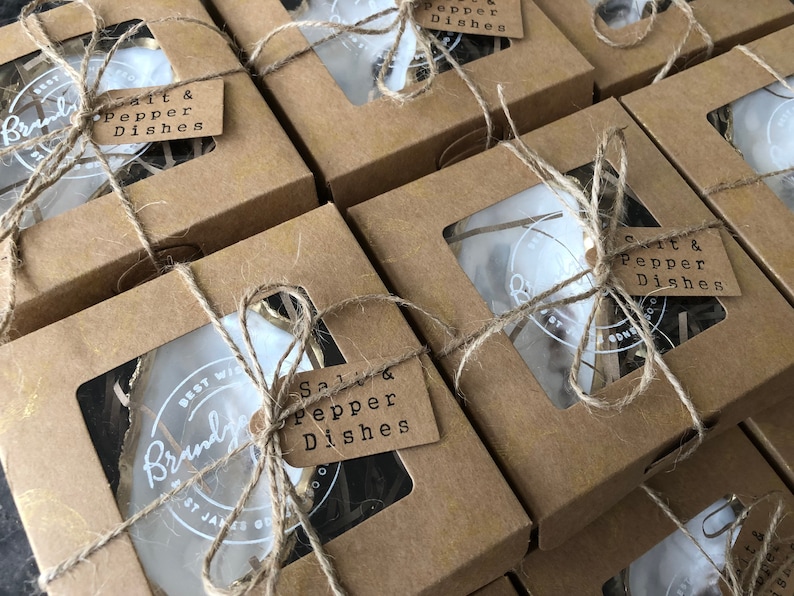 Op de afbeelding: Meerdere bruine kartonnen dozen met doorzichtige ramen, elk met een witte keramische zout- en peperstrooierset. De dozen zijn vastgebonden met touw en hebben een bruin papieren etiket met de tekst "Salt & Pepper Dishes".