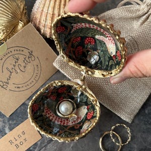 May include: Two seashell ring boxes with gold edging, one open and one closed, displaying a ring. The interior has a bird and berry pattern. A small card with the text "Best Wishes From Brandyark Cottage" is visible.