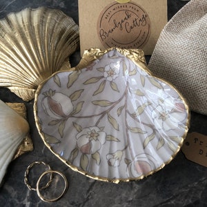 May include: A set of three decorative trinket dishes shaped like seashells. The dishes are made of white and gold painted resin. The largest dish has a floral pattern and a gold rim. The smaller dishes are solid gold. The dishes are displayed on a grey surface with a brown tag that reads "Trinket Dish".