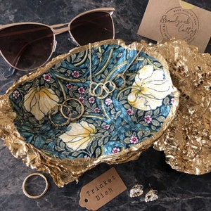 May include: A gold trinket dish with a floral pattern and a gold, textured rim. The dish is filled with jewelry, including a necklace, a ring, and earrings. The dish is sitting on a dark gray surface.