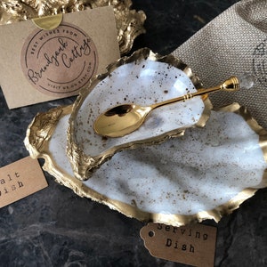 May include: Two white ceramic oyster shell dishes with gold accents. The smaller dish is filled with a gold spoon. The larger dish has a brown tag that says "Serving Dish". The smaller dish has a brown tag that says "Salt Dish".