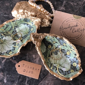 May include: Two decorative trinket dishes made from oyster shells, featuring a floral pattern in green, white, and yellow. The edges are trimmed with gold. A small tag reads "Trinket Dish". A card with the text "Best Wishes From Brandjork Cottage" is also present.