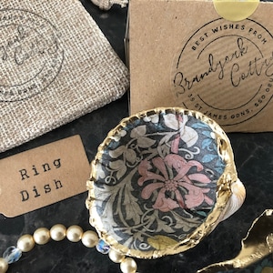 May include: A small, gold-rimmed, ceramic ring dish with a floral pattern. The dish is sitting on a dark surface. The text "Ring Dish" is printed on a brown tag. The text "Best Wishes From Brandyark Cottage 38 St James Gdns, S502BU" is printed on a brown box.