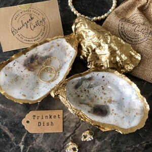 May include: Two gold-rimmed oyster shell trinket dishes, one with a white interior and the other with a white interior with gold flecks. The dishes are on a dark grey surface with a brown tag that reads "Trinket Dish".