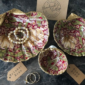 May include: Three decorative ceramic trinket dishes shaped like seashells. The dishes are decorated with a red, green and gold floral pattern. The largest dish contains a pearl necklace. The smaller dishes are labelled "Ring Dish" and "Trinket Dish".