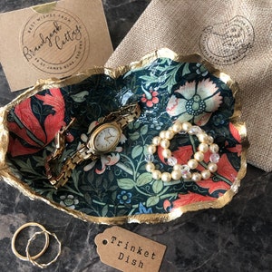 Pu&ograve; includere: Un portagioie decorativo con un motivo floreale rosso, rosa e blu, bordato d'oro. Il piatto contiene un orologio d'oro, un braccialetto di perle e un braccialetto a catena d'oro. Sono presenti anche un piccolo sacchetto e un'etichetta con la scritta "Trinket Dish".