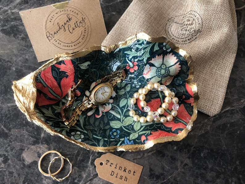 Pu&ograve; includere: Un portagioie decorativo con un motivo floreale rosso, rosa e blu, bordato d'oro. Il piatto contiene un orologio d'oro, un braccialetto di perle e un braccialetto a catena d'oro. Sono presenti anche un piccolo sacchetto e un'etichetta con la scritta "Trinket Dish".