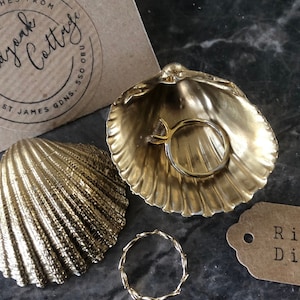 May include: Two gold-colored seashell ring dishes. One dish has a ring inside. The other dish is next to a small gold-colored ring. A brown tag with the text "Ring Dish" is in the lower right corner of the image.