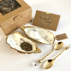 Op de afbeelding: Een set van twee gouden gemarmerde oesterschelp zout- en peperbakjes met twee gouden lepels met kristallen handvatten. De bakjes zijn gevuld met zout en peper en worden geleverd in een bruine doos met een label waarop "Brandywine Cottage" staat.