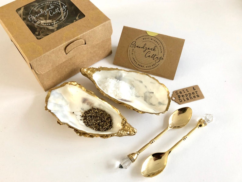 Op de afbeelding: Een set van twee gouden gemarmerde oesterschelp zout- en peperbakjes met twee gouden lepels met kristallen handvatten. De bakjes zijn gevuld met zout en peper en worden geleverd in een bruine doos met een label waarop "Brandywine Cottage" staat.