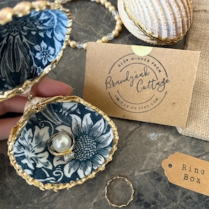 May include: A ring box made from two seashells with a gold trim. The inside of the box is lined with a blue and white floral fabric. A pearl ring is sitting inside the box. The box is sitting on a gray surface with a brown tag that says "Ring Box".