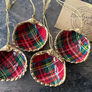 May include: Four seashell ornaments with a red, green, and yellow plaid pattern. The edges are trimmed with gold, and each has a gold string for hanging. A small card with text is in the background.