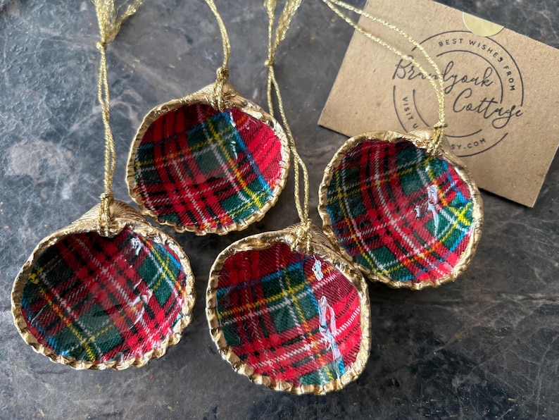 May include: Four seashell ornaments with a red, green, and yellow plaid pattern. The edges are trimmed with gold, and each has a gold string for hanging. A small card with text is in the background.