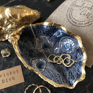 May include: Gold oyster shell trinket dish with a blue and white peacock feather design. The dish is labelled "Trinket Dish".