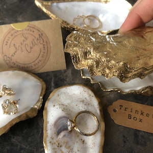 May include: Three gold-rimmed oyster shell trinket dishes. One dish has a pearl necklace and earrings, another has a ring, and the third has a tag that says "Trinket Box".