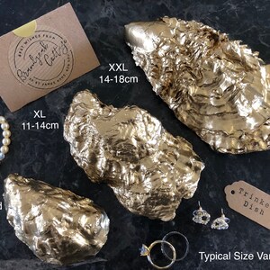 Pu&ograve; includere: Piatti portagioie a forma di conchiglia di ostrica dipinti d'oro, in varie misure: Standard (9-11 cm), XL (11-14 cm) e XXL (14-18 cm). L'immagine include un braccialetto di perle, orecchini e anelli. Un'etichetta recita "Trinket Dish".