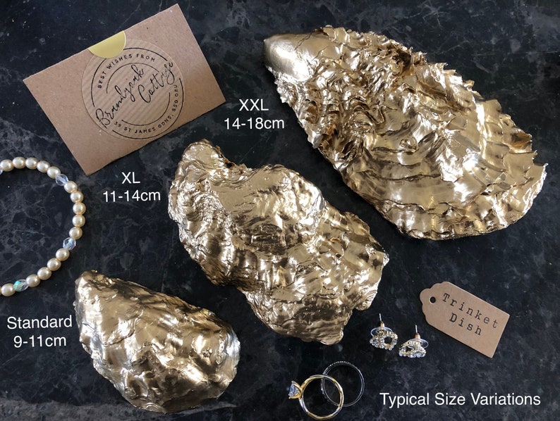 Pu&ograve; includere: Piatti portagioie a forma di conchiglia di ostrica dipinti d'oro, in varie misure: Standard (9-11 cm), XL (11-14 cm) e XXL (14-18 cm). L'immagine include un braccialetto di perle, orecchini e anelli. Un'etichetta recita "Trinket Dish".