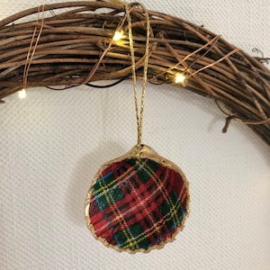 May include: A Christmas ornament made from a seashell, featuring a red, green, and yellow plaid pattern. The shell's edge is trimmed with gold paint, and it hangs from a gold string. The ornament is set against a rustic wreath.