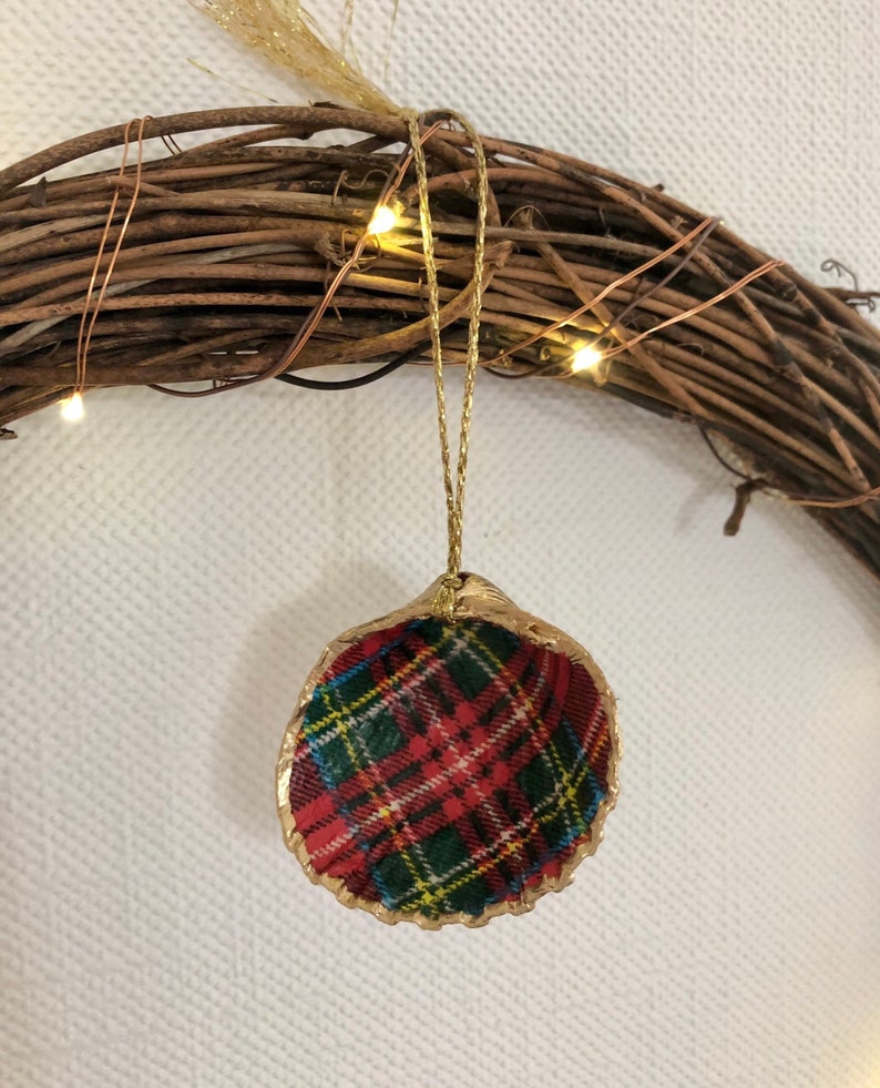 May include: A Christmas ornament made from a seashell, featuring a red, green, and yellow plaid pattern. The shell's edge is trimmed with gold paint, and it hangs from a gold string. The ornament is set against a rustic wreath.