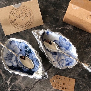 Könnte beinhalten: Zwei weiße Keramik-Salz- und Pfefferstreuer in Form von Austernmuscheln mit blauen Blumenmustern und silbernen Löffeln. Die Löffel haben Kristallgriffe. Der Text "Salt & Pepper Dishes" befindet sich auf einem braunen Etikett.