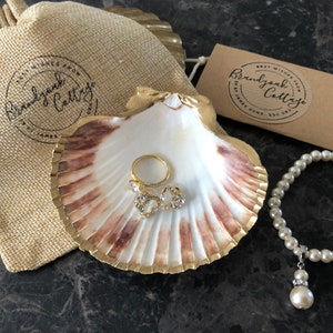 Puede incluir: Una concha blanca con un borde dorado, utilizada como joyero, con un anillo de oro y dos peque&ntilde;os pendientes de oro con piedras transparentes en su interior. Una cadena de perlas blancas con un colgante de perla y cristal cuelga junto a la concha. Una bolsa de papel marr&oacute;n con el texto "Brandywine Cottage" est&aacute; en el fondo.