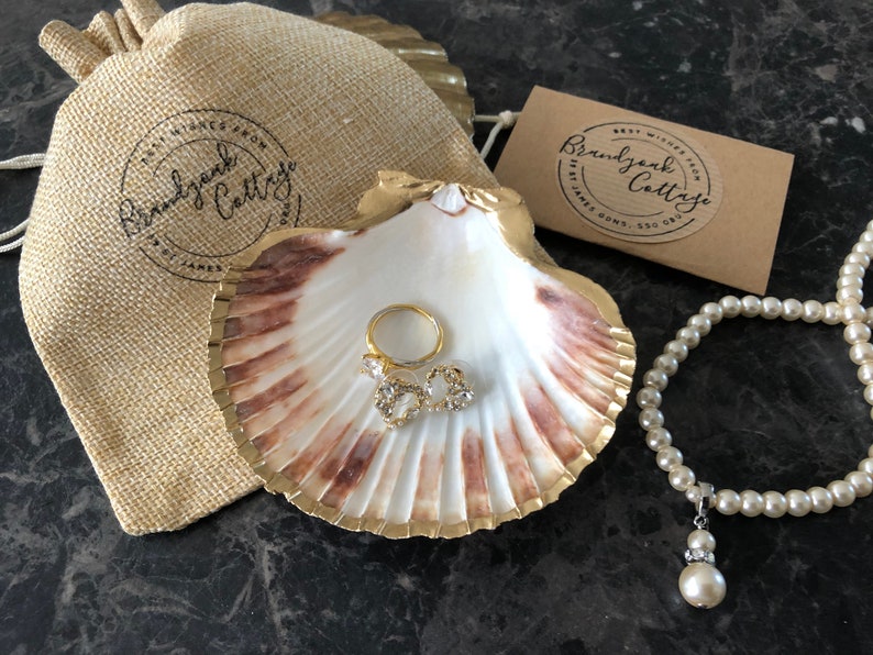 Puede incluir: Una concha blanca con un borde dorado, utilizada como joyero, con un anillo de oro y dos peque&ntilde;os pendientes de oro con piedras transparentes en su interior. Una cadena de perlas blancas con un colgante de perla y cristal cuelga junto a la concha. Una bolsa de papel marr&oacute;n con el texto "Brandywine Cottage" est&aacute; en el fondo.